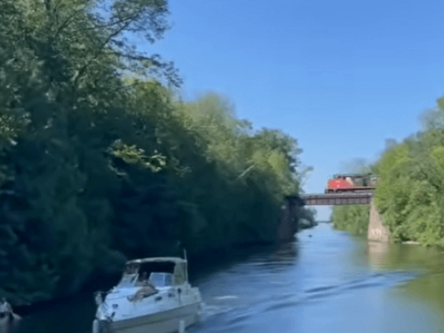 Boomershines cruising narrow canal with canoe, other boat, and trains passing on bridge